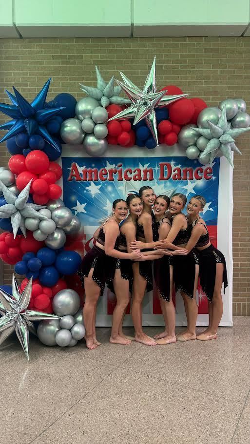 Seven women in black dresses pose for a photo in front of a backdrop with balloons and stars.