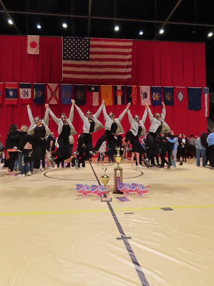 Group of dancers jumping and holding trophies in a gymnasium. Flags from different countries and a red backdrop are present.