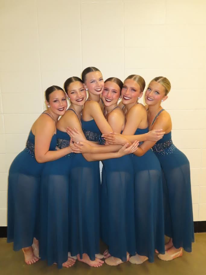 Five women in blue dresses stand together, smiling, and hugging each other in a room with white walls.