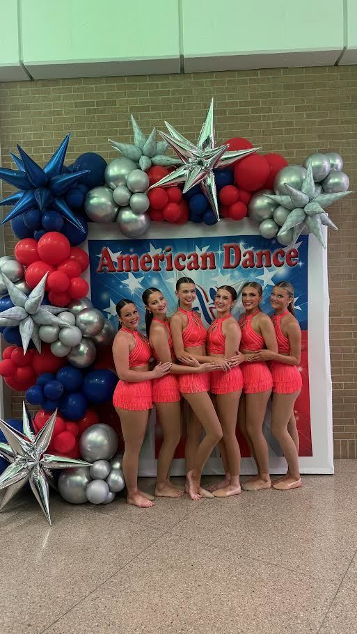 Six women in red outfits stand next to each other in front of a sign that says "American Dance."