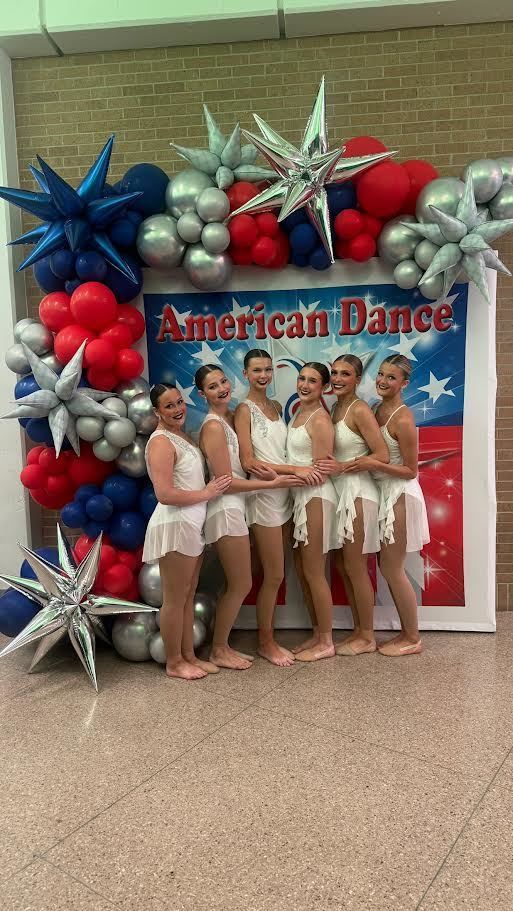 Five women wearing white dresses stand together in front of a backdrop reading "American Dance" with balloons and stars.