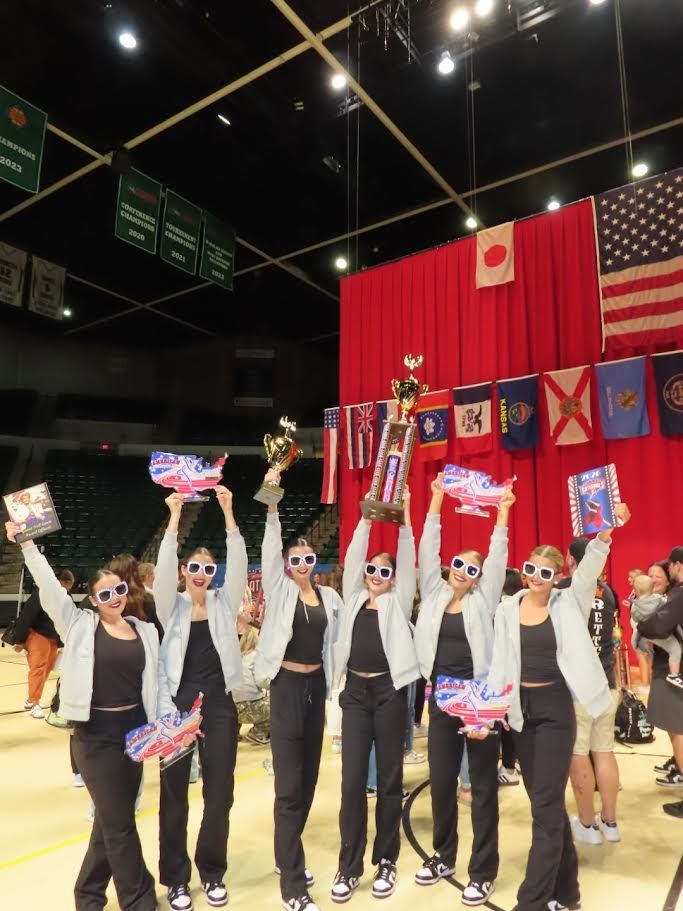 A group of eight individuals in matching attire hold trophies and flags, standing on a stage. A red curtain and flags in the background.