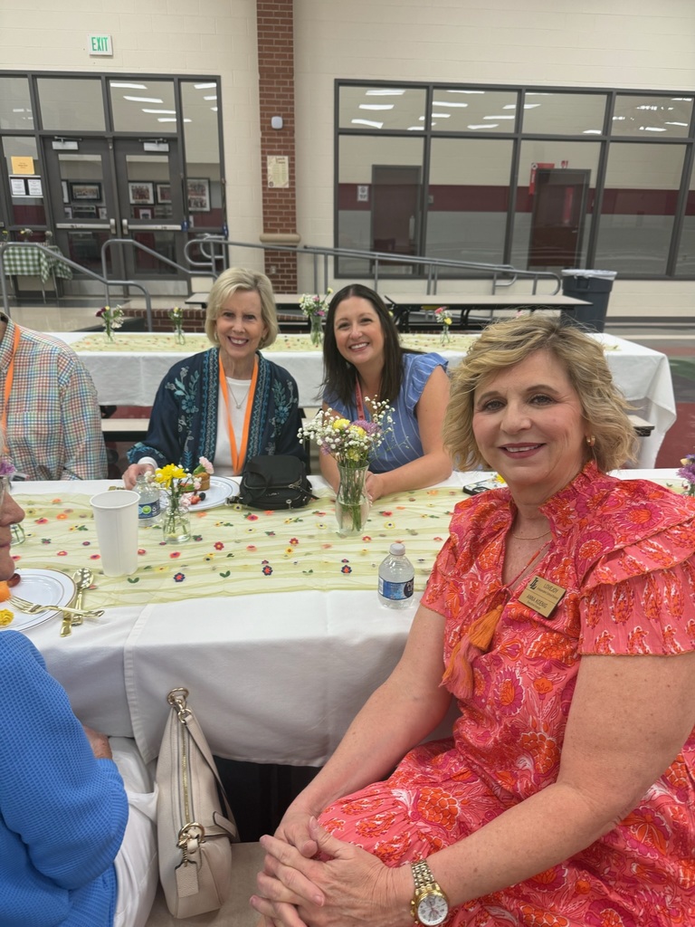 Four women in dresses sit at a table with flowers and plates, with two women smiling at the camera.