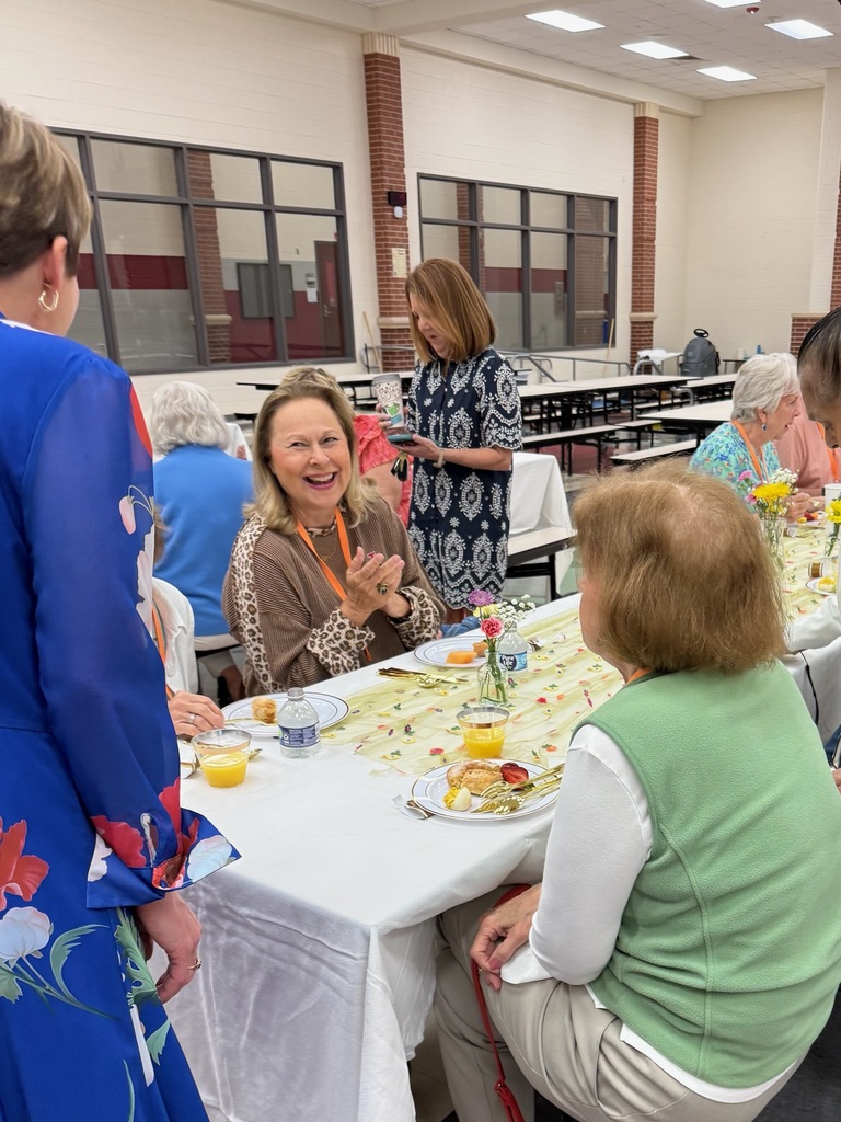 People sit at a table with plates, cups, and flowers. One woman smiles while others talk.