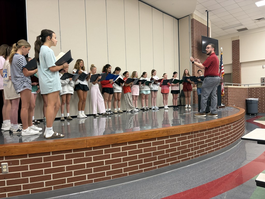 A choir practices on a stage, led by a man in red, with students in various colors holding music books.