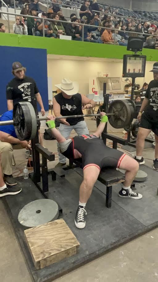 A man bench presses heavy weights with three spotters, while an audience watches from the stands.