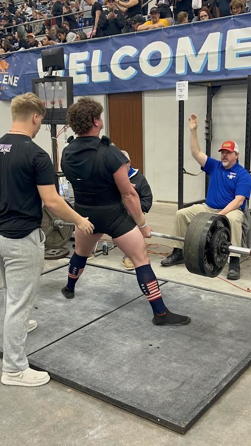 A man squats while lifting a barbell, wearing a black singlet and socks with a flag pattern. A judge signals with raised hands. Spectators are in the background.