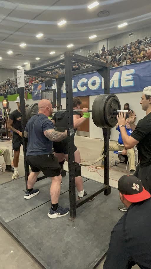 A man performs a squat with a barbell in a gym. Several people observe from behind and on the side.