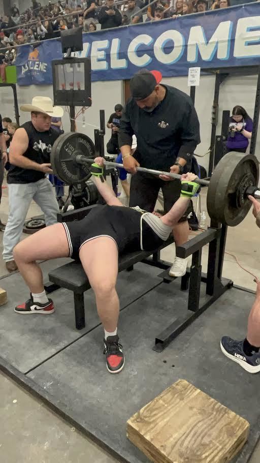 An athlete performs a bench press with a barbell while a spotter assists, wearing a hat, gloves, and sneakers.
