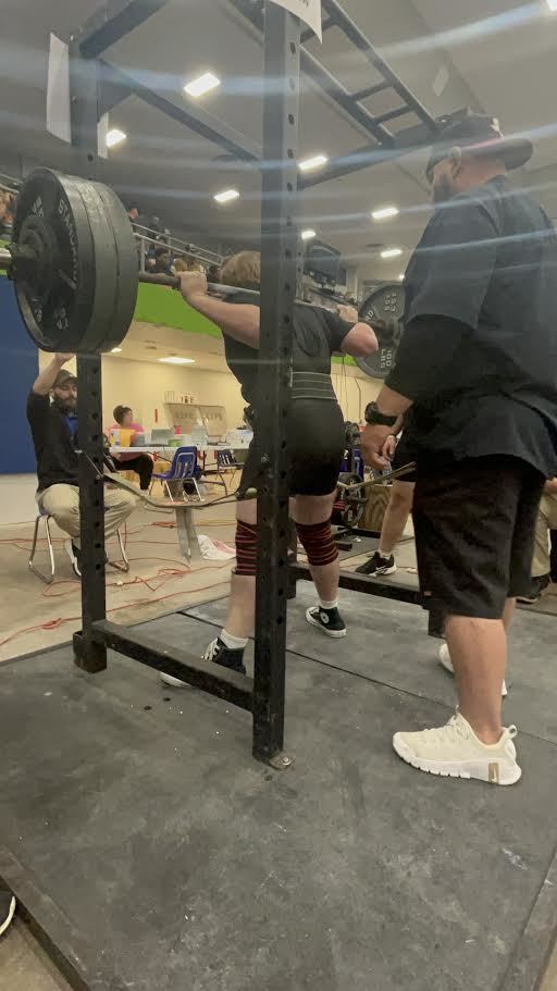 A man squats under a barbell with assistance from another person in a gym with other people in the background.