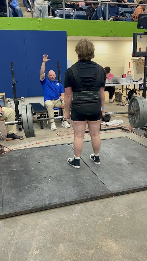 An older man stands on a platform, lifting a barbell with a spotter nearby. The gym features a blue wall, and others are present in the background.