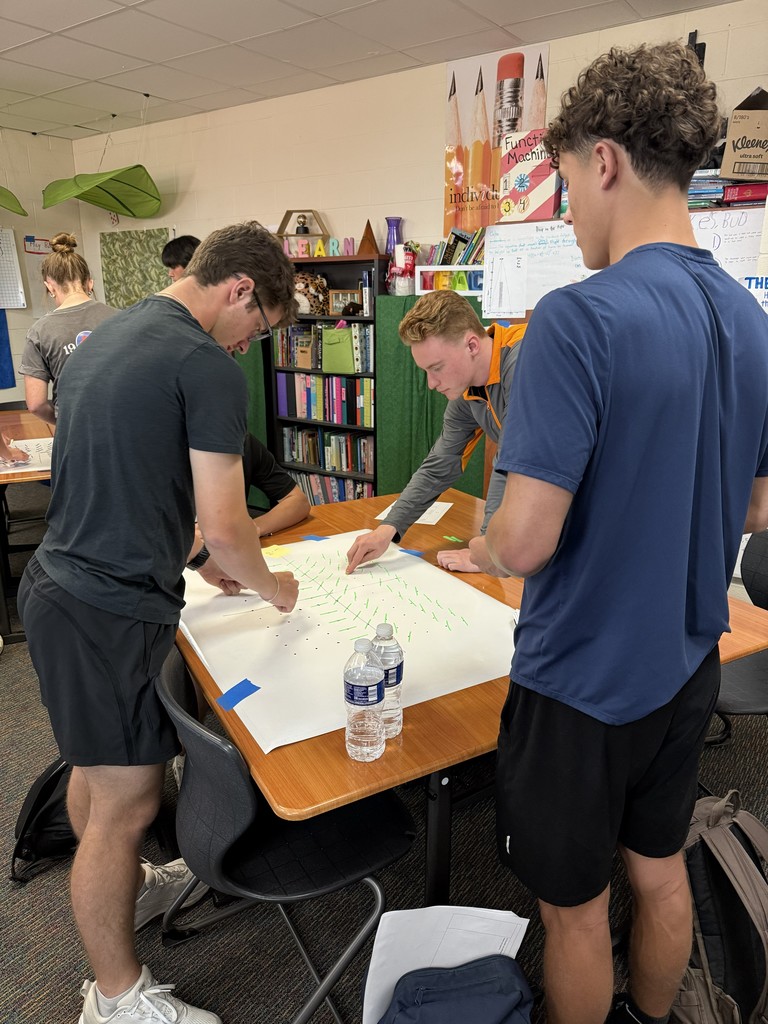 Three young people examine a large sheet of paper on a desk. One person writes with a marker.