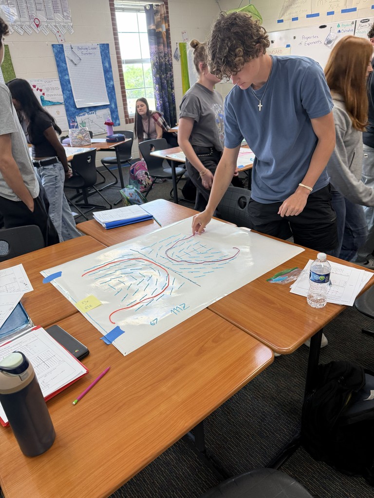 A student writes on a large sheet of paper on a table in a classroom. Others stand nearby.