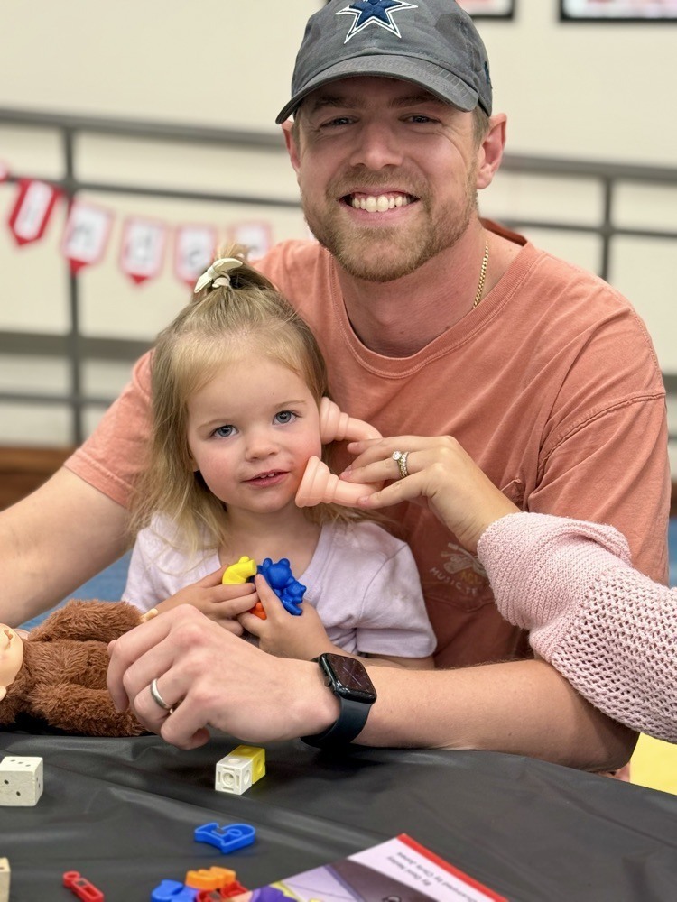 A smiling man and a young girl sit at a table with toys. The man wears a baseball cap and ring.