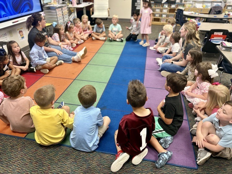A classroom with children sitting on the floor in a circle. A teacher stands in front of them.