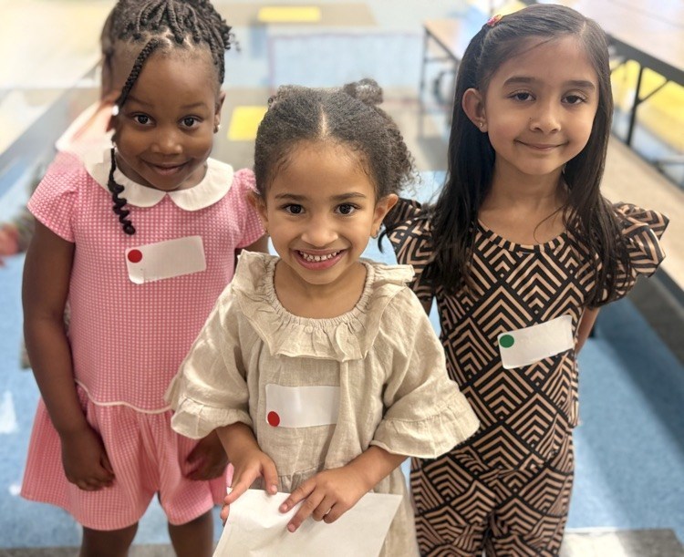 Three young girls in school uniforms stand together, smiling, in a hallway with a bench in the background.