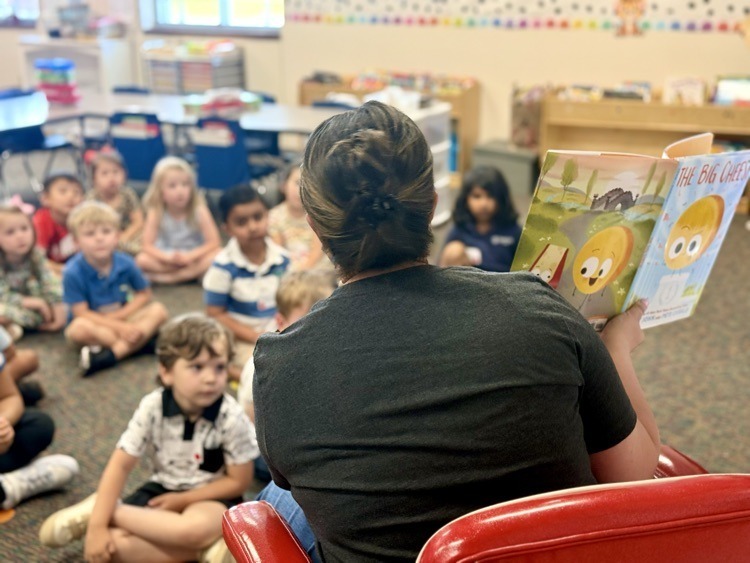 A person reads a book to children sitting on the floor. Some children wear sneakers, others wear sandals.
