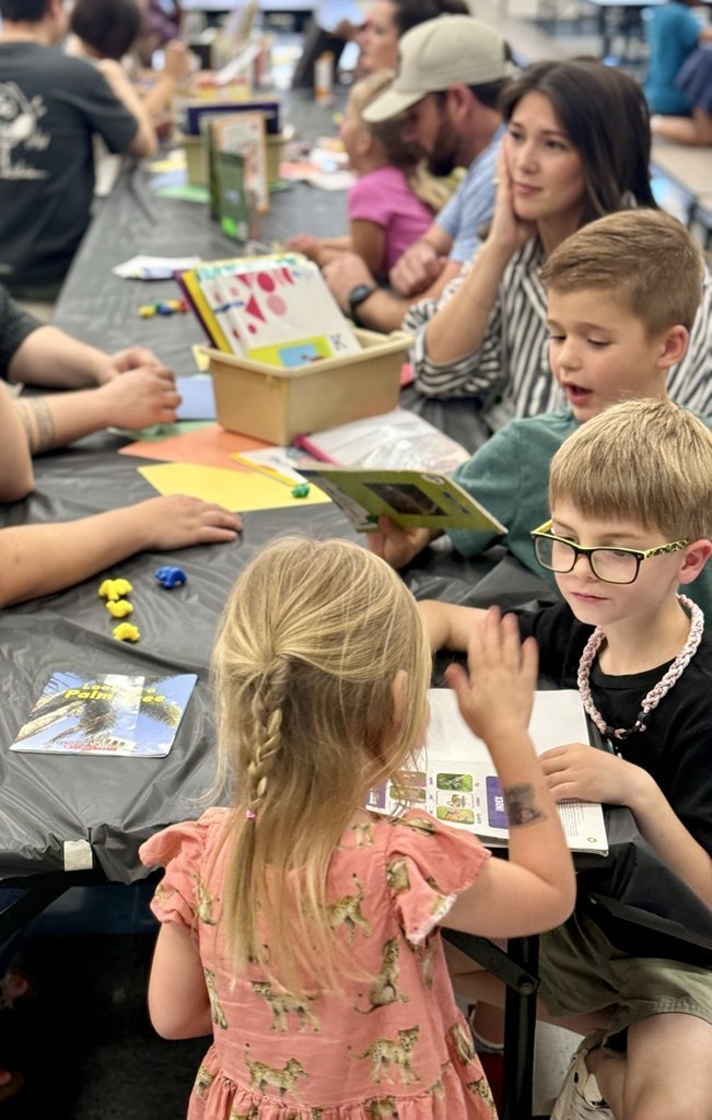 A group of children and adults at a table with books, craft materials, and toys.