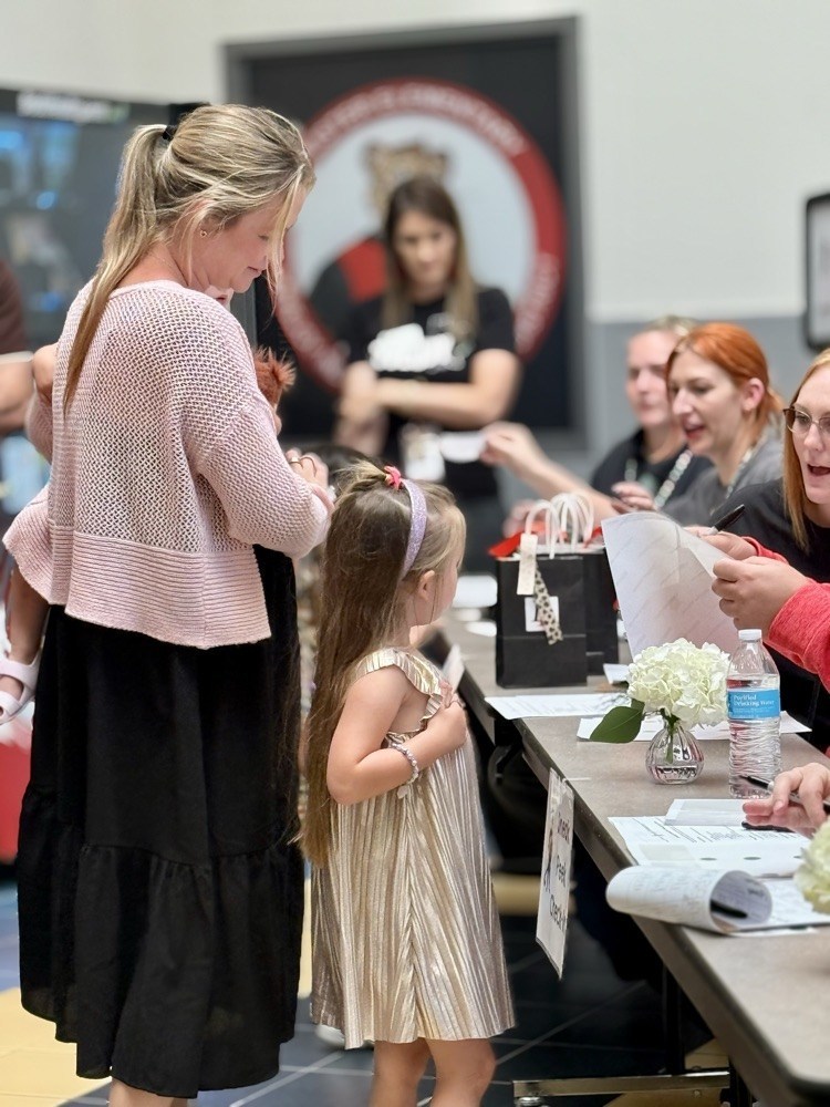 A woman and young girl stand at a table. The woman wears a pink sweater, and the girl a gold dress.