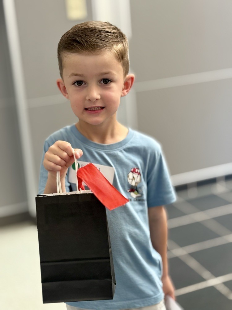 Boy holding a black paper bag with red ribbon, wearing a blue shirt and white shorts, standing in a tiled room.