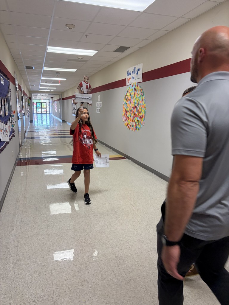 A young child in red is walking and looking at an adult in gray inside a school hallway.