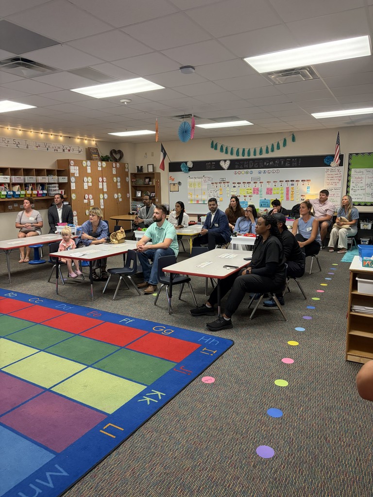 A classroom with tables and chairs, a colorful rug, and a group of people sitting and standing.