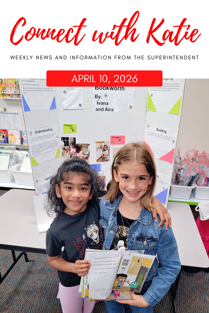 Two girls pose in a classroom, one holding a book. They stand before a display with posters, text, and photos.