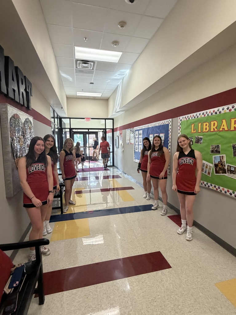 Group of girls in cheerleading uniforms stands in a hallway with a library sign on the wall.