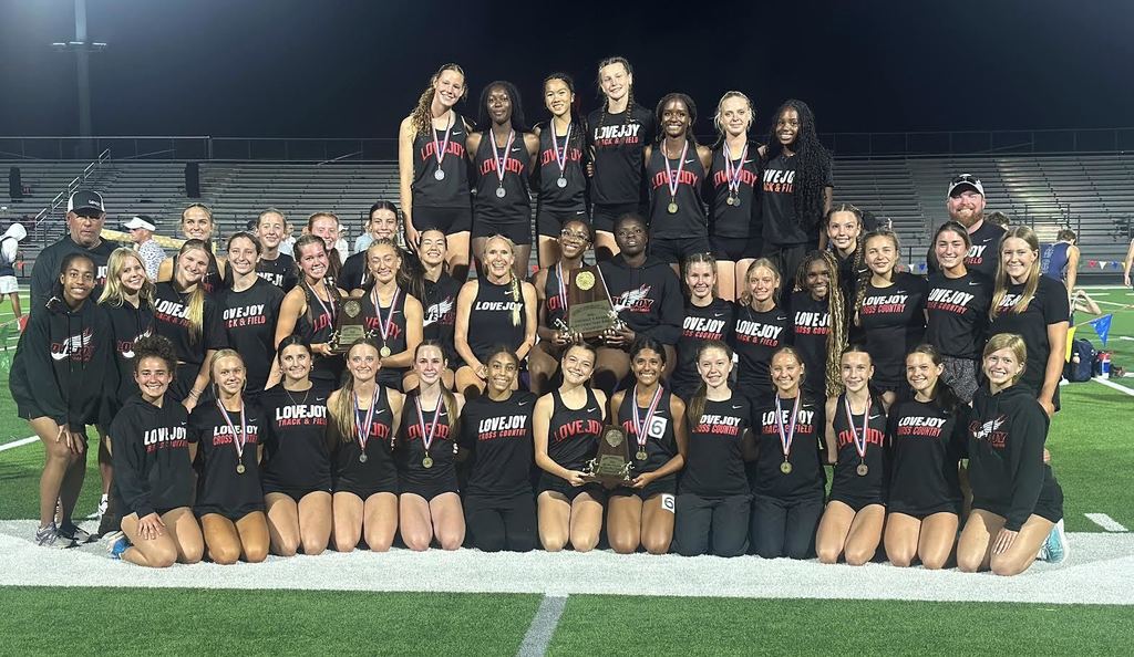 A team wearing black uniforms and medals poses on a field with trophies, stadium lights in background.