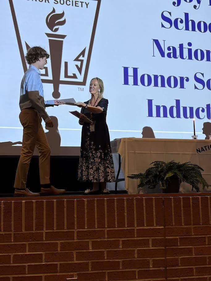 A person hands a paper to a woman on stage. Behind them, a screen displays the words "National Honor Society Induction".