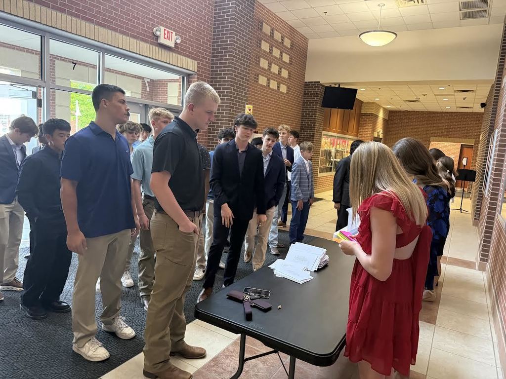 Group of people standing around a table in a hallway. One person wearing a red dress is reading.