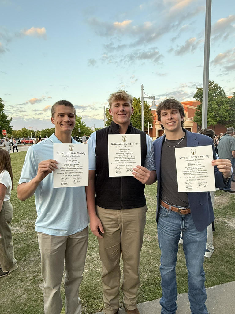 Three people in light clothing stand on grass holding diplomas. One wears a gray jacket. Trees and buildings are in the background.
