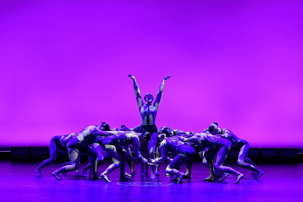 Dancers in dark blue costumes perform on stage, with one dancer raised above a circle of others. Purple lights illuminate the background.