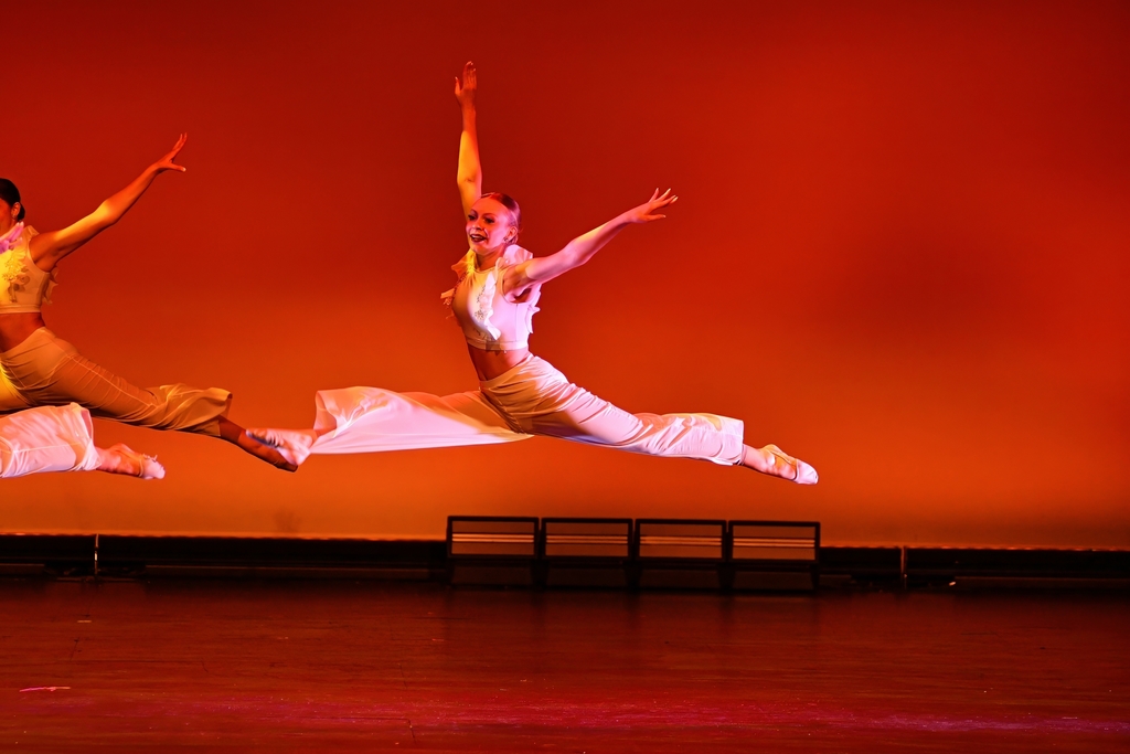 Two dancers leap on stage, arms and legs extended, against a red backdrop, in front of a dark stage.