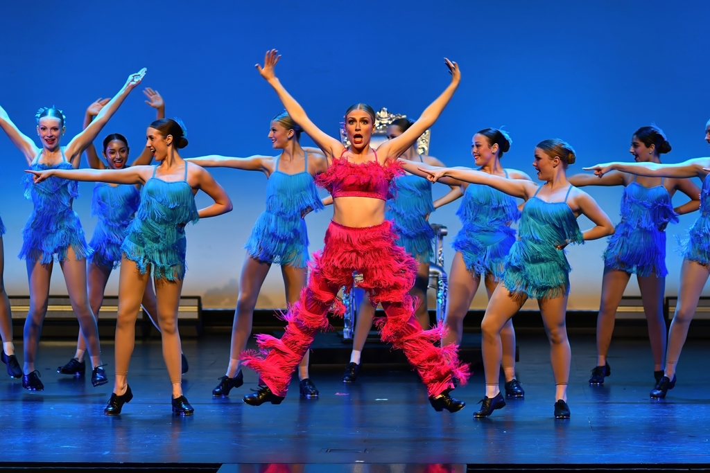 Dancers perform on stage; one in pink feathers stands out, all wear blue and pink costumes, and black shoes.