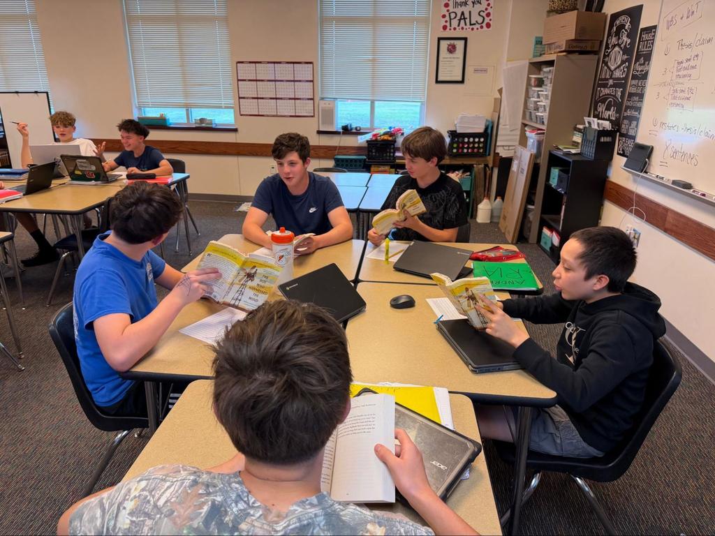Several students sit at a table, reviewing books and papers. A whiteboard and cabinets are in the background.