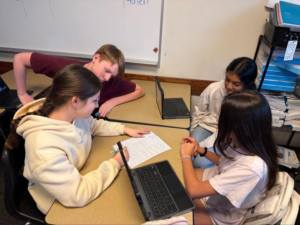 Four students in a classroom, two at a desk, two leaning over another desk, all looking at a laptop.
