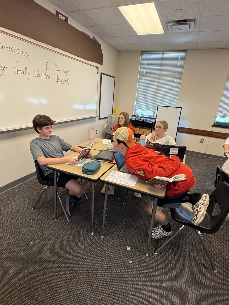 Five people sit at desks in a classroom, three of them are using laptops. The room has a whiteboard on the wall.