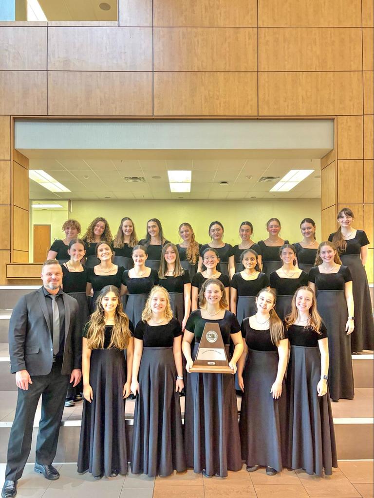 A choir of women in black dresses, posing with a trophy in a hall with wooden paneled walls.