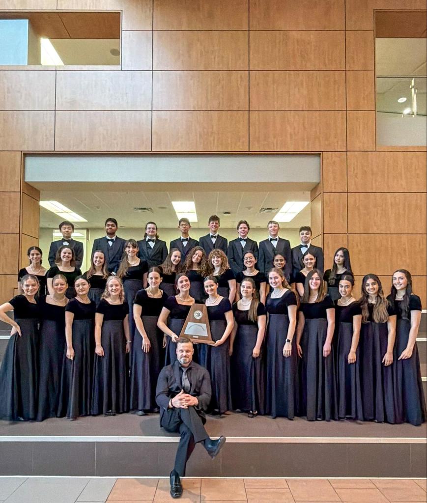Choir members in black dresses stand on a stage, posing for a group photo. A trophy is held by some members. Behind them, a wooden wall.