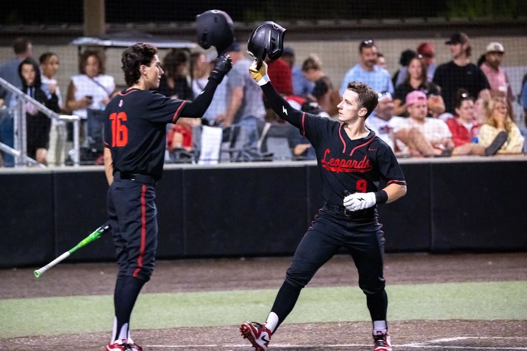 Two baseball players celebrate with raised helmets. Player 16 stands left, holding a bat. Player 55 wears a red hat. Spectators watch from behind a fence.