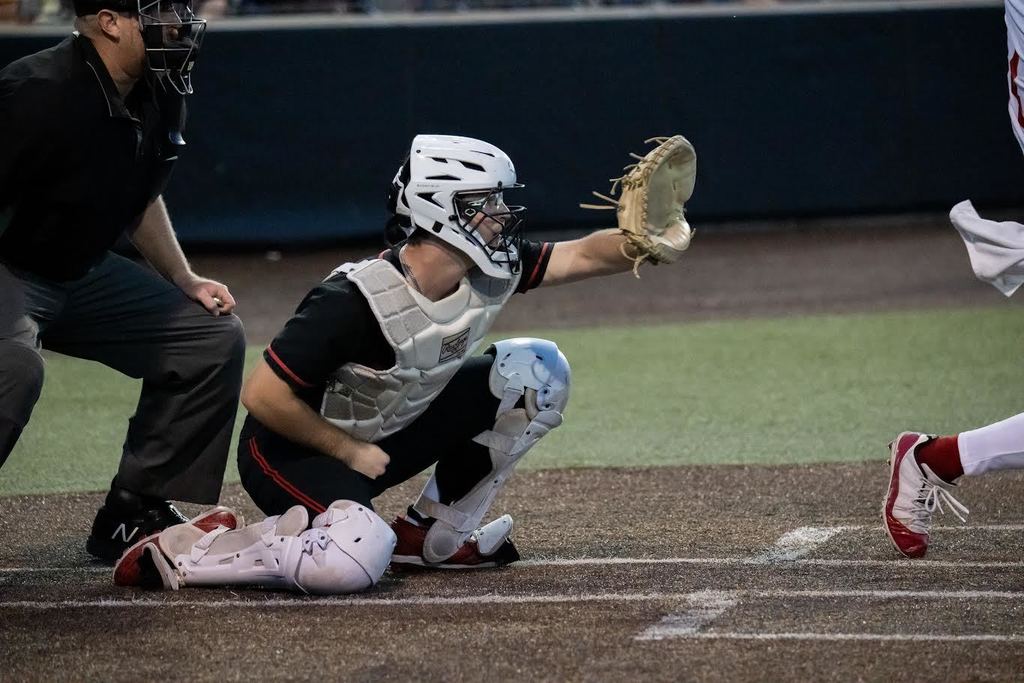 A baseball player in a crouch catches a ball while a catcher stands behind. A referee watches from the side.