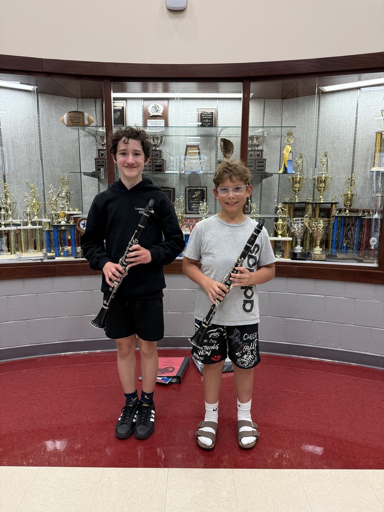 Two boys stand, holding clarinets, in a room with red flooring and a trophy-filled display case.