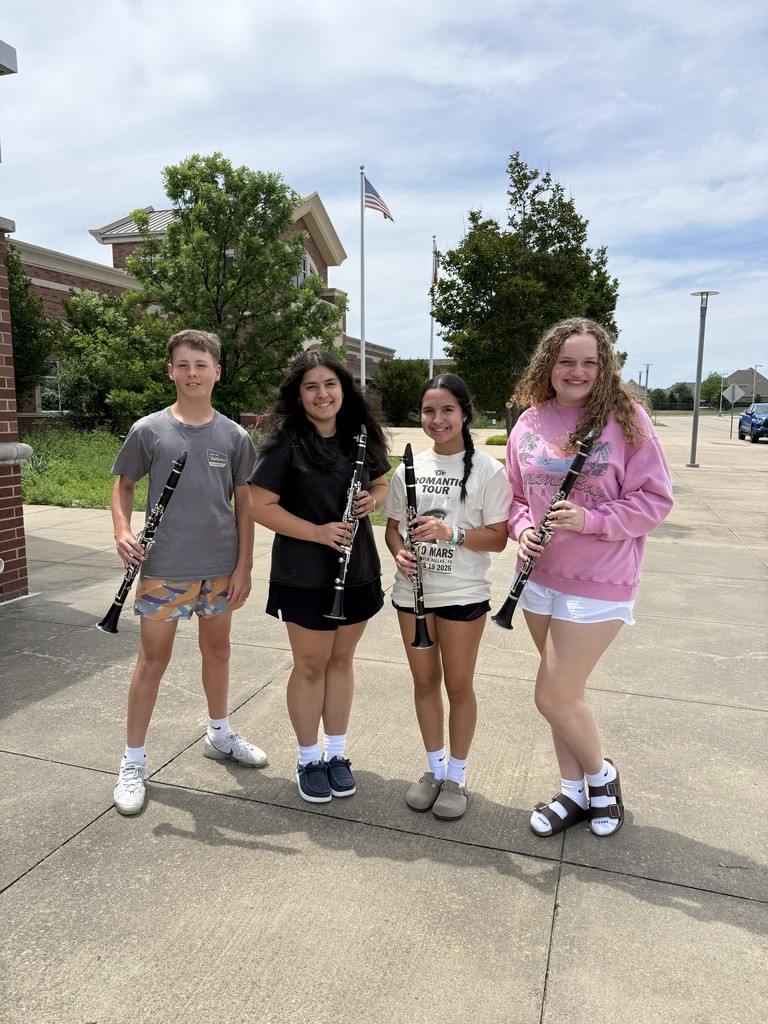 Four young individuals, each holding a clarinet, pose in front of a building with trees and a flag.