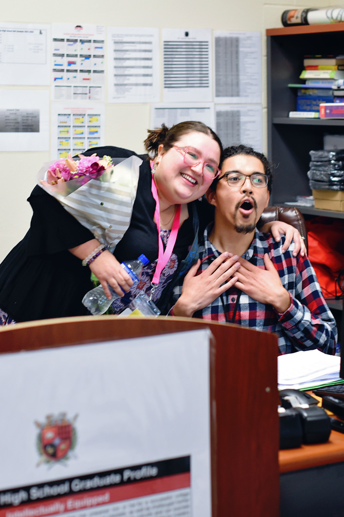 Two people in a room with a desk; a woman wearing glasses and a scarf around her neck.