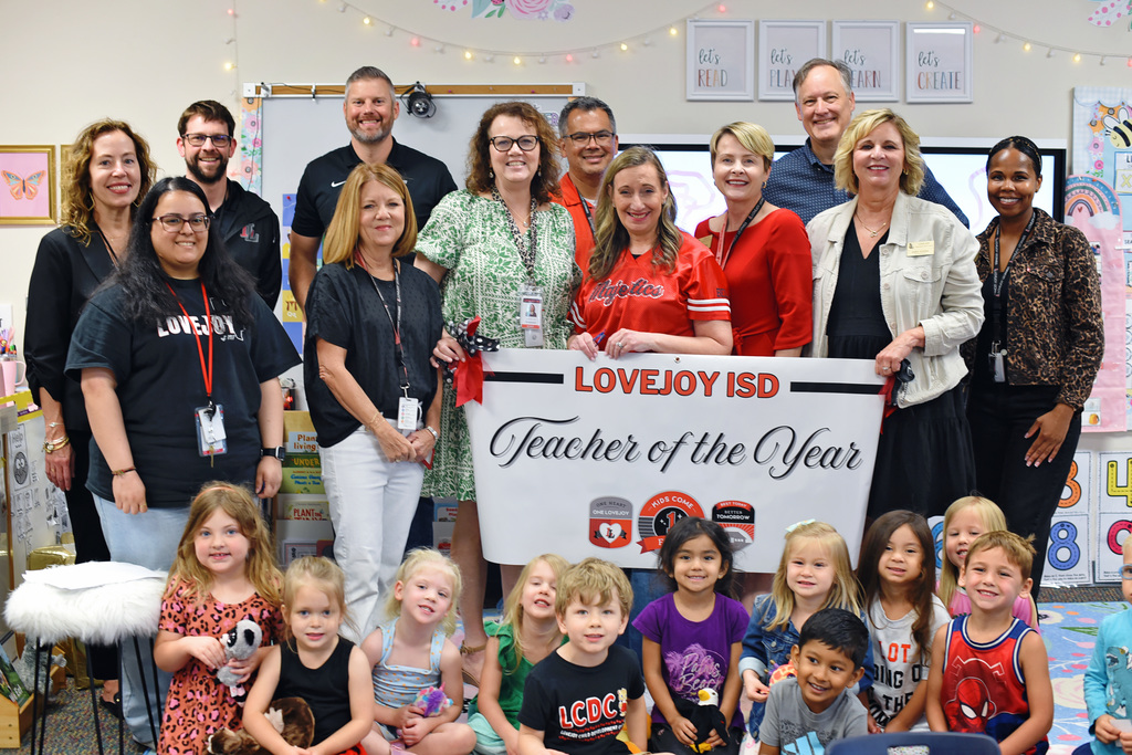 Group of adults and children in a classroom. One woman holds a sign that reads "Teacher of the Year".