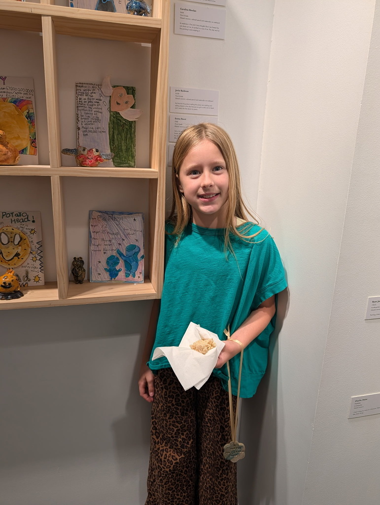 A young girl smiles, holding a sandwich, in front of a white wall. Shelves with art pieces are behind her.