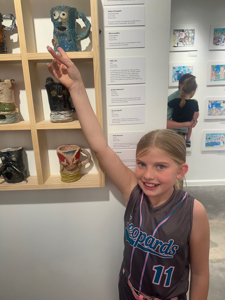 A girl in a sports jersey points at a tall mug displayed on a shelf in an art gallery.