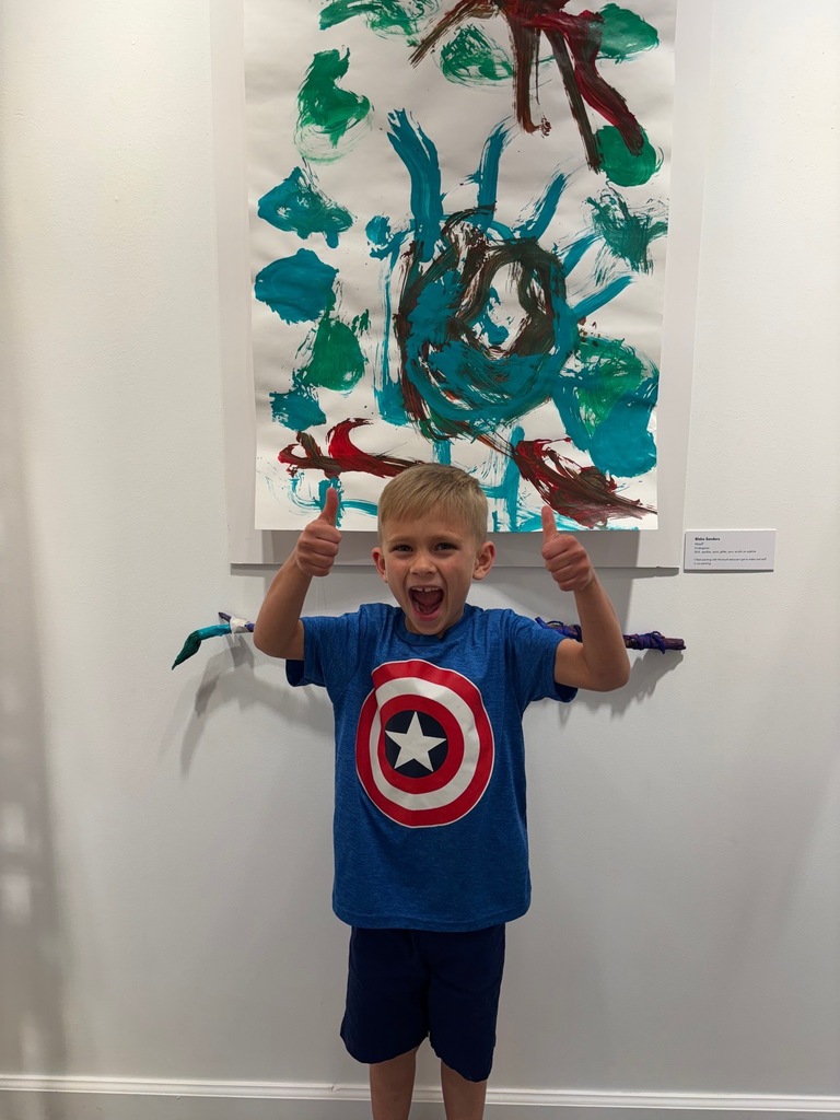Young boy wearing blue shirt and shorts stands in front of colorful artwork on a white wall.