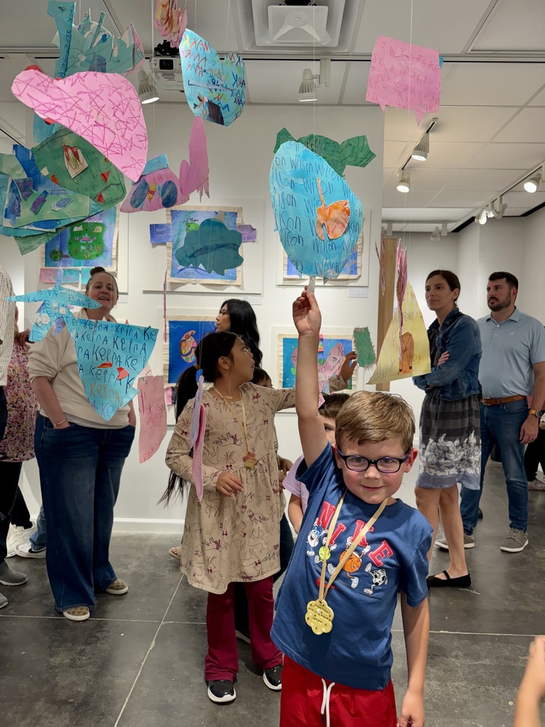 Several children and adults in a gallery. A boy raises his hand. Colorful artwork hangs from ceiling.